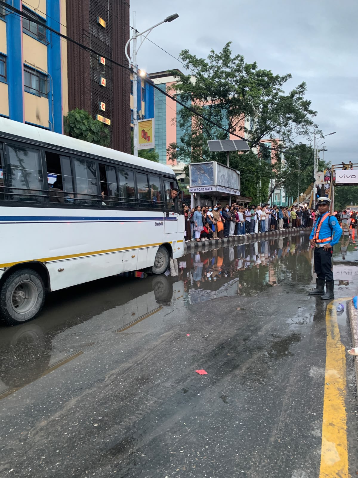 Crowd waiting on the kerb beside a flooded bus lay-by at Newroad Gate, with a traffic police officer standing nearby